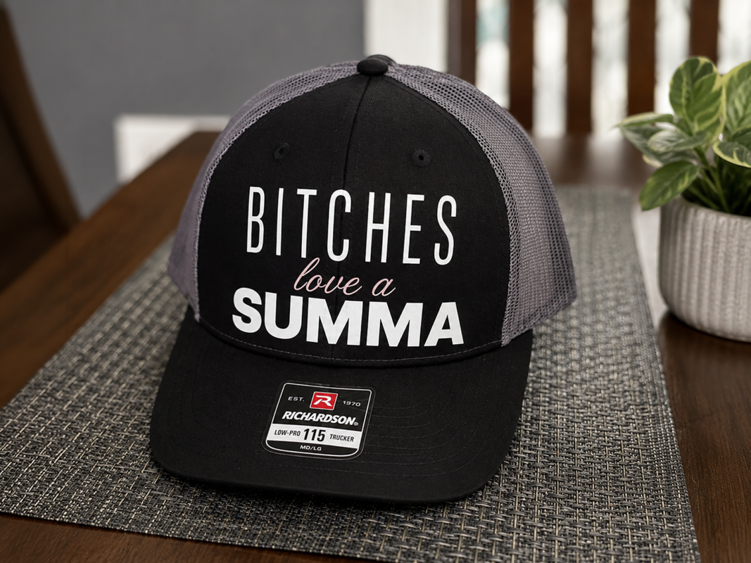 Bitches love a Summa trucker hat in black and gray with a bold phrase design, shown on a tabletop.