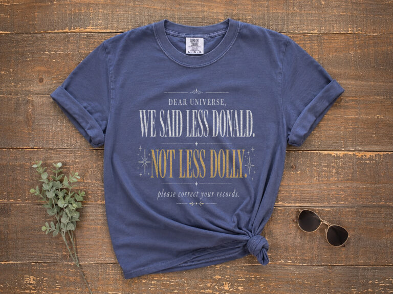 Front view of a garment-dyed unisex tee featuring a white-and-gold text satire graphic; a less Donald more Dolly shirt.