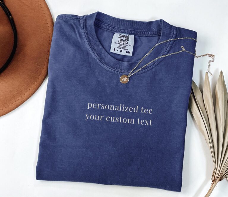 Folded blue personalized Comfort Colors shirt with sample custom text in a flat-lay arrangement.