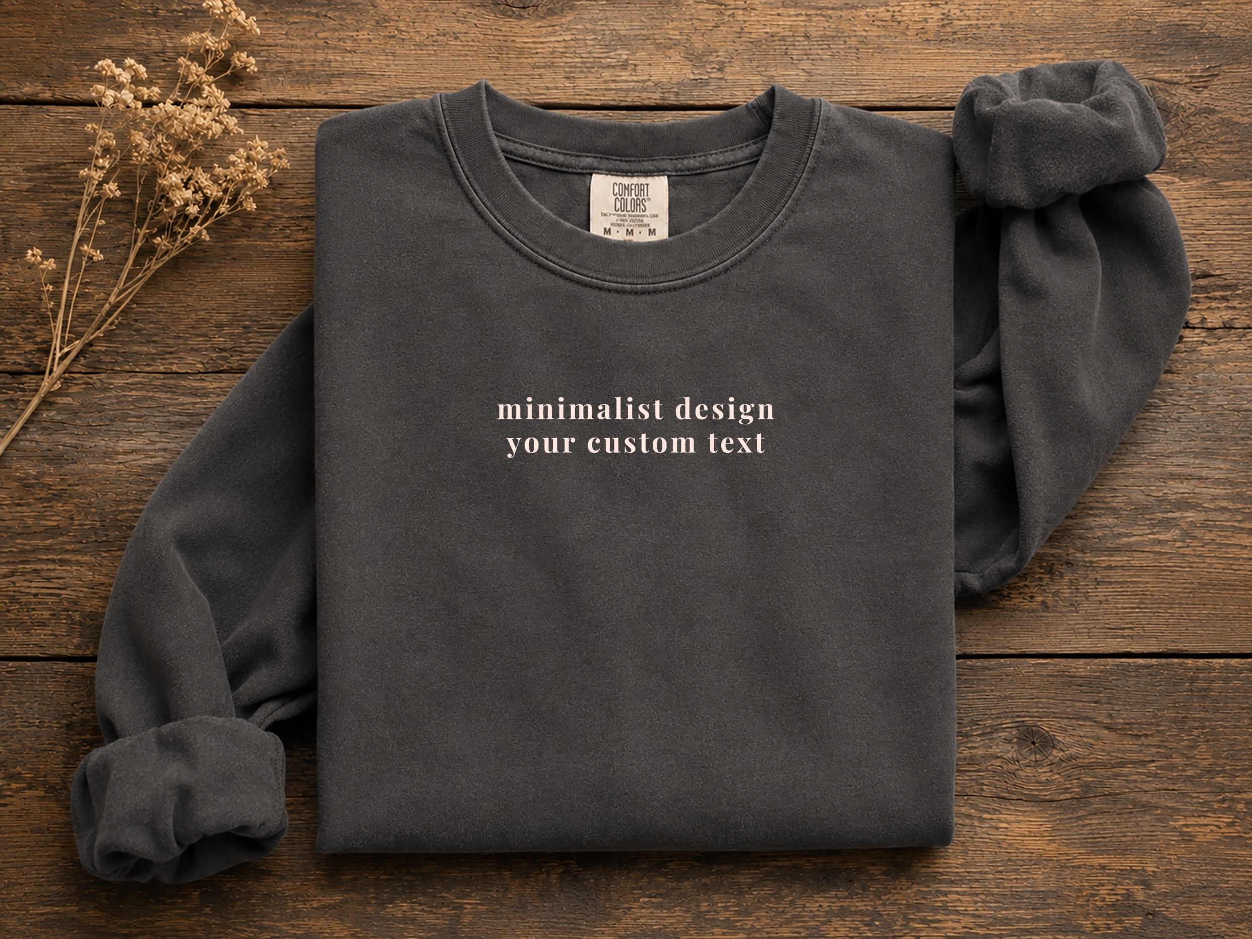 Folded personalized text sweatshirt mockup on a wooden surface with dried floral stems.